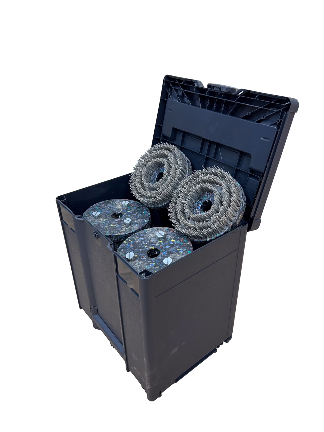 Un robuste conteneur de rangement en plastique noir avec son couvercle à charnière ouvert, révélant quatre grandes têtes d'outil circulaires à l'intérieur. Deux des pièces sont des roues de brosse abrasives denses avec des poils métalliques en anneaux concentriques ; les deux autres sont des tampons épais de polissage en matériau composite multicolore recyclé, fixés par des vis métalliques visibles. Le conteneur présente des éraflures et des traces d'usure légères ; la photo est prise sur un fond blanc propre avec des ombres douces, mettant en valeur la texture des poils de la brosse et du matériau recyclé. Les objets semblent être des têtes de rechange pour des appareils industriels de nettoyage de sols ou de préparation de surfaces.